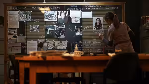 A detective writes on a crime board covered with photos maps and suspect notes