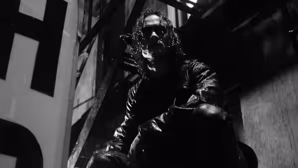 A long-haired man in dark clothes crouches beneath a metal staircase