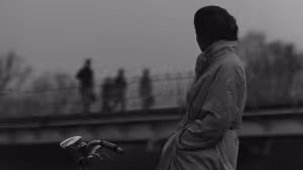 A woman in a coat stands beside a riverside railing, facing distant figures