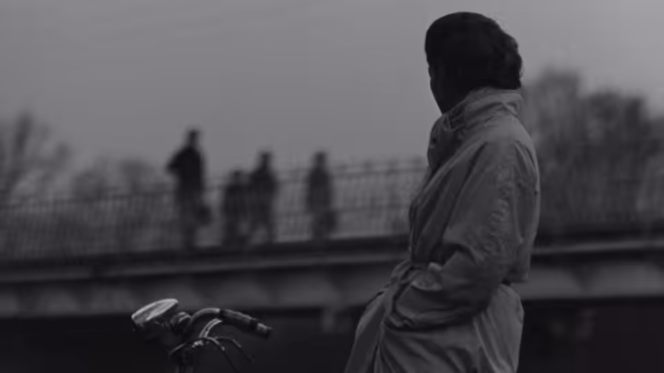 A woman in a coat stands beside a riverside railing, facing distant figures