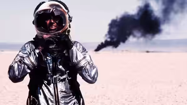 Astronaut in silver suit adjusts straps in desert with smoke rising behind