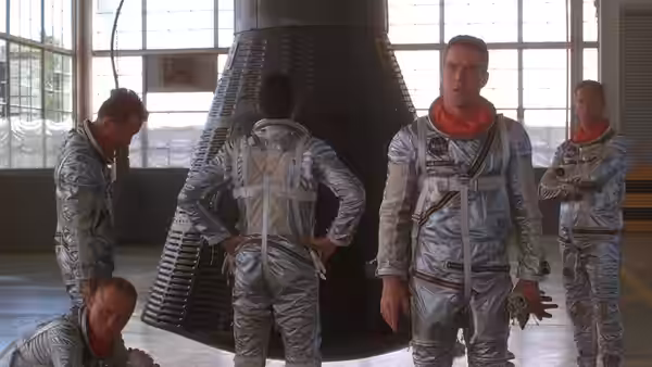 Astronauts in silver suits gather around a large rocket nozzle indoors