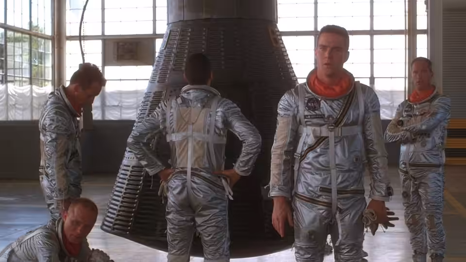 Astronauts in silver suits gather around a large rocket nozzle indoors
