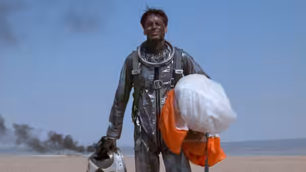 Astronaut in damaged suit walks across desert carrying helmet and parachute