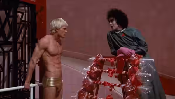Rocky Horror (Peter Hinwood) faces Dr. Frank-N-Furter (Tim Curry) beside red ribbons and a cage
