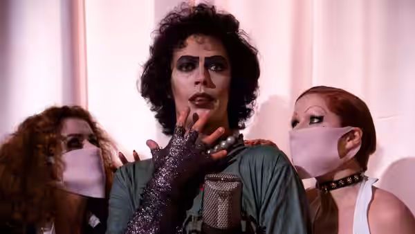 Dr. Frank-N-Furter (Tim Curry) stands between two masked attendants in white face coverings