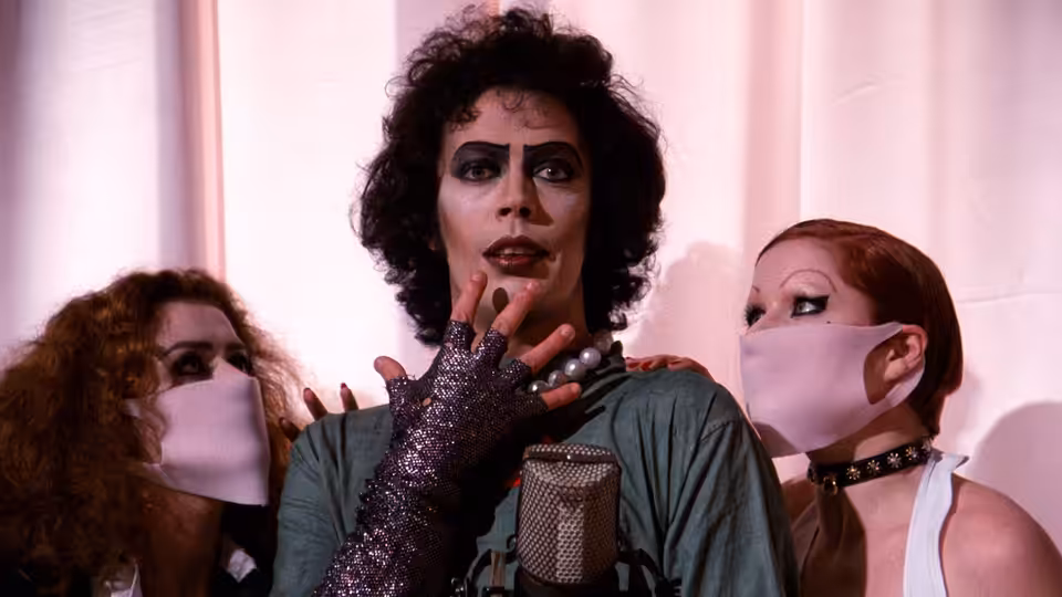 Dr. Frank-N-Furter (Tim Curry) stands between two masked attendants in white face coverings