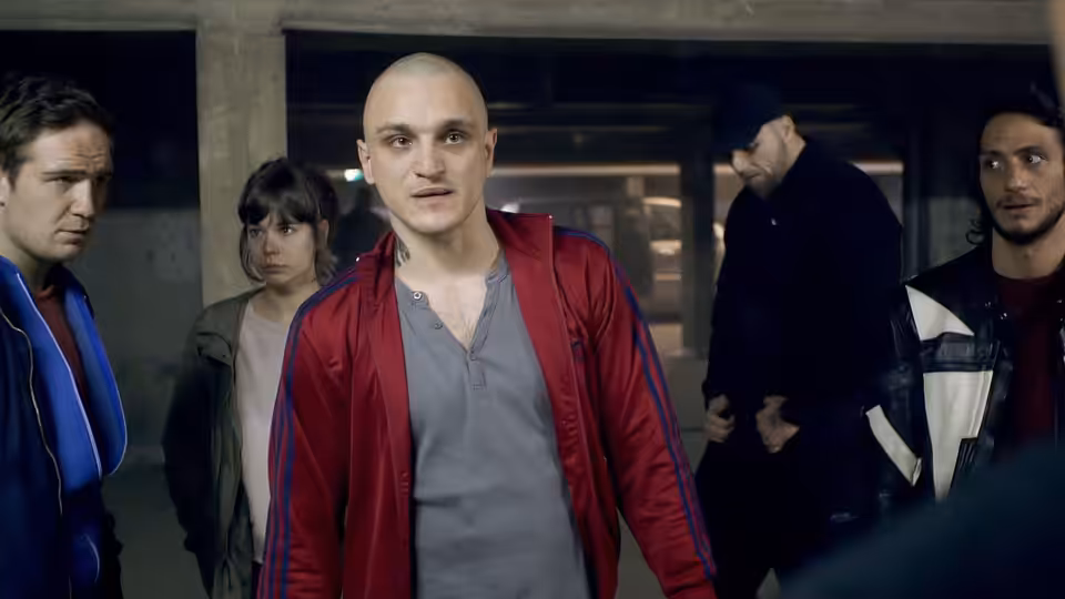 A man in a red jacket stands facing forward with others behind him