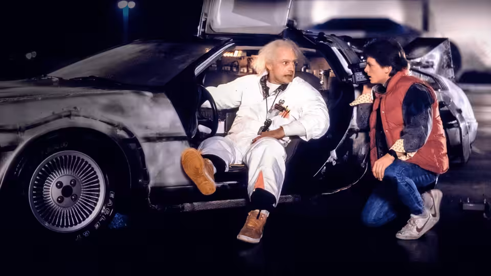 Emmett Brown (Christopher Lloyd) seated in the DeLorean as Marty McFly (Michael J. Fox) kneels beside