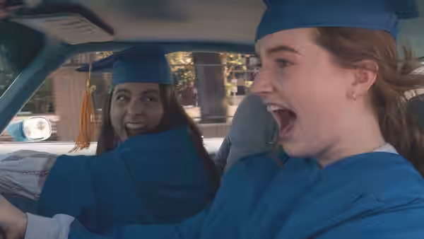 Molly (Beanie Feldstein) drives a car in graduation gown mouth open while Amy (Kaitlyn Dever) looks back