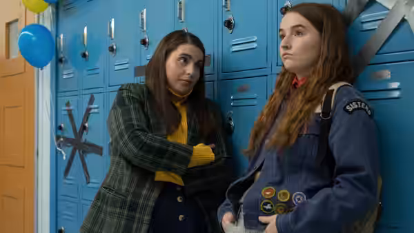 Molly (Beanie Feldstein) stands arms crossed beside Amy (Kaitlyn Dever) at blue school lockers