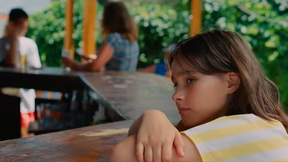 Sophie (Frankie Corio) rests her arms on a wooden table in a shaded outdoor café