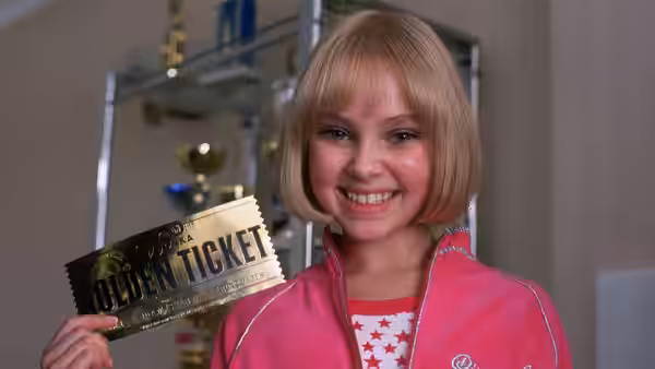 A girl smiles while holding up a golden ticket in a trophy room