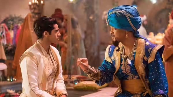 Aladdin (Mena Massoud) seated with the Genie (Will Smith) at a market stall