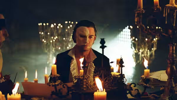 The Phantom seated at a table surrounded by lit candles and papers