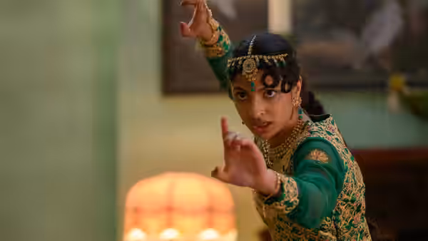 Ria Khan (Priya Kansara) in green and gold raises both hands in a room