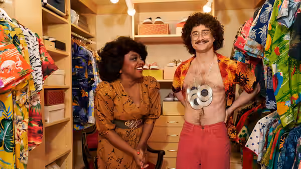 Weird Al (Daniel Radcliffe) with open shirt and CDs taped to chest stands beside a woman in a closet
