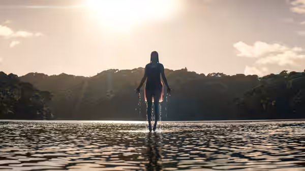 A woman in a red dress walking into shallow water towards the sun