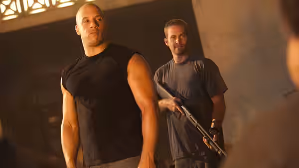 Dominic Toretto (Vin Diesel) stands beside Brian O'Conner (Paul Walker), who holds a rifle indoors