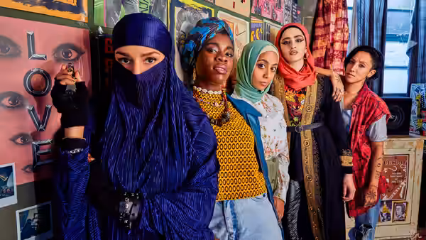 Five women in hijabs and niqab standing side by side against a poster-covered wall