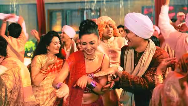 A bride in red dances with wedding guests beside a smiling man in a white turban
