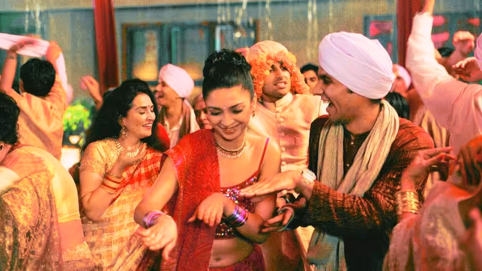 A bride in red dances with wedding guests beside a smiling man in a white turban