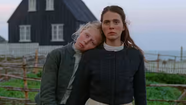 Two women stand close together in front of a dark house near the sea