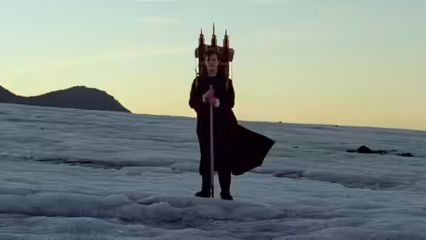 A man holding a staff stands on snow with a large wooden structure on his back
