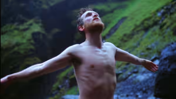 A shirtless bearded man standing with arms outstretched in a rocky green valley