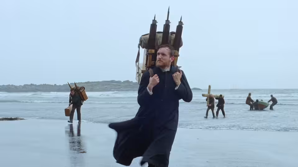 A man carrying a wooden church structure running across a wet beach with others following
