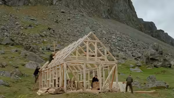 Workers assembling the wooden frame of a building on a rocky green hillside