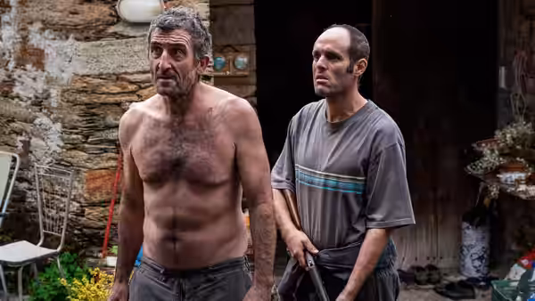 Two men stand outside a stone building one holding a tool