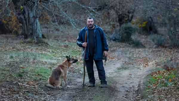 A bearded man stands with a dog on a wooded dirt path