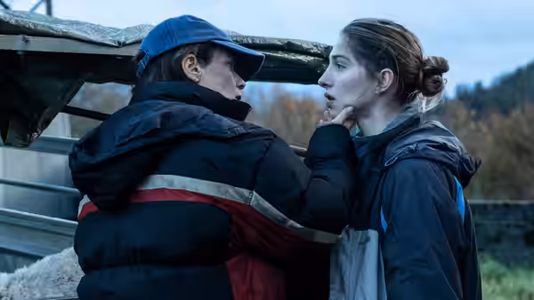 Two women stand face to face outdoors with one touching the chin