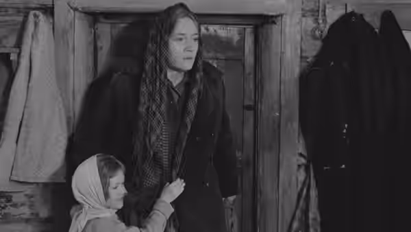 A woman stands indoors as a child clings to her coat