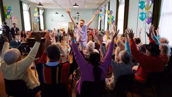 Man in hospital gown raises arms among elderly people with hands up in hall