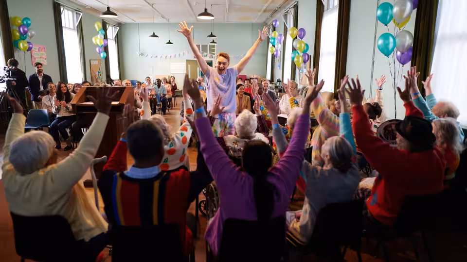 Man in hospital gown raises arms among elderly people with hands up in hall