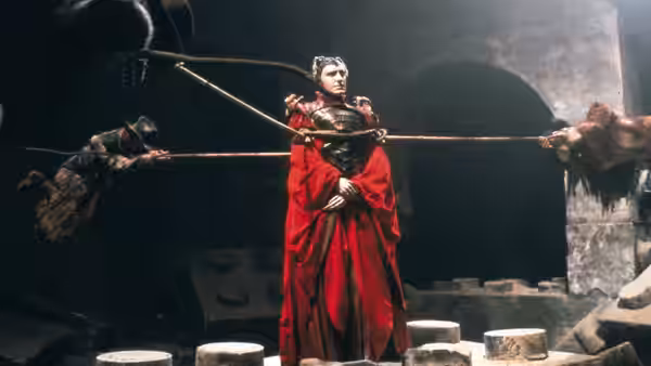 A woman in a red gown stands bound by spears in a stone chamber