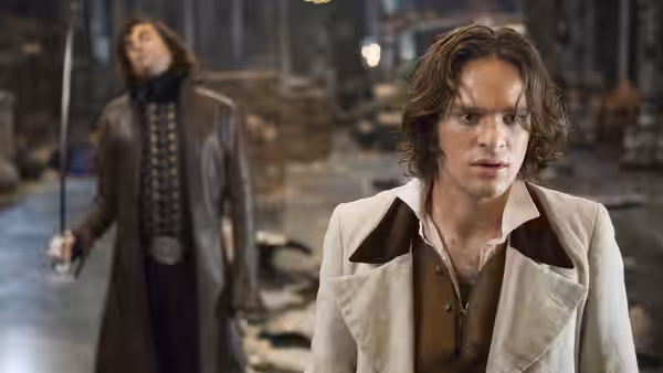 Tristan looks ahead in a crowded room as a long-haired man stands behind him holding a sword