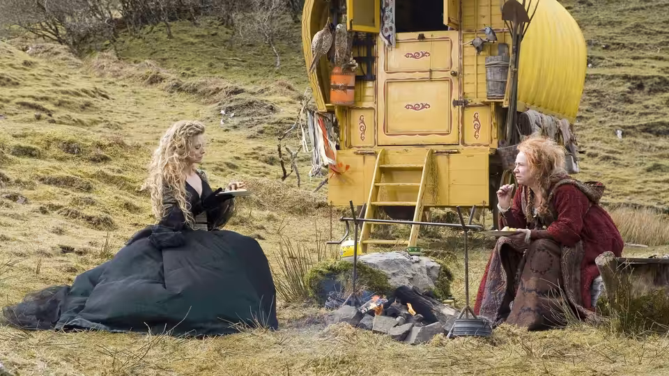 A blonde woman and a red-haired traveler sit beside a yellow caravan on a grassy hillside