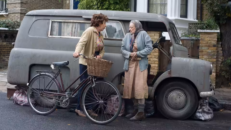 Alan Bennett (Alex Jennings) speaks with Miss Shepherd (Maggie Smith) beside her van and a bicycle