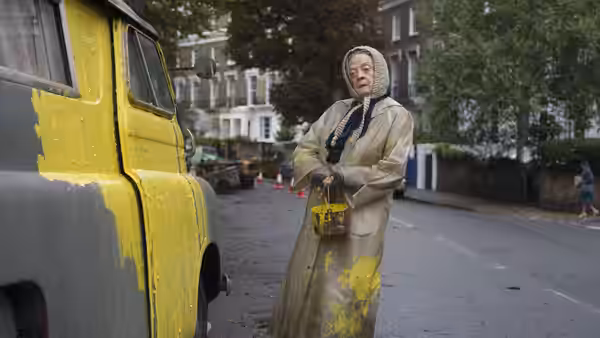 Miss Shepherd (Maggie Smith) stands beside her yellow-painted van, holding a paintbrush