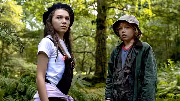 A girl and boy standing among trees in a dense green forest