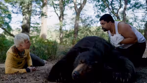 A blonde woman lying beside a bear while a man kneels over it