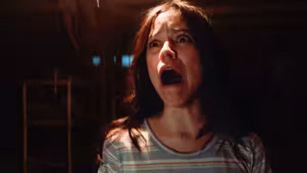 A woman with dark hair screams with mouth open in a dark room