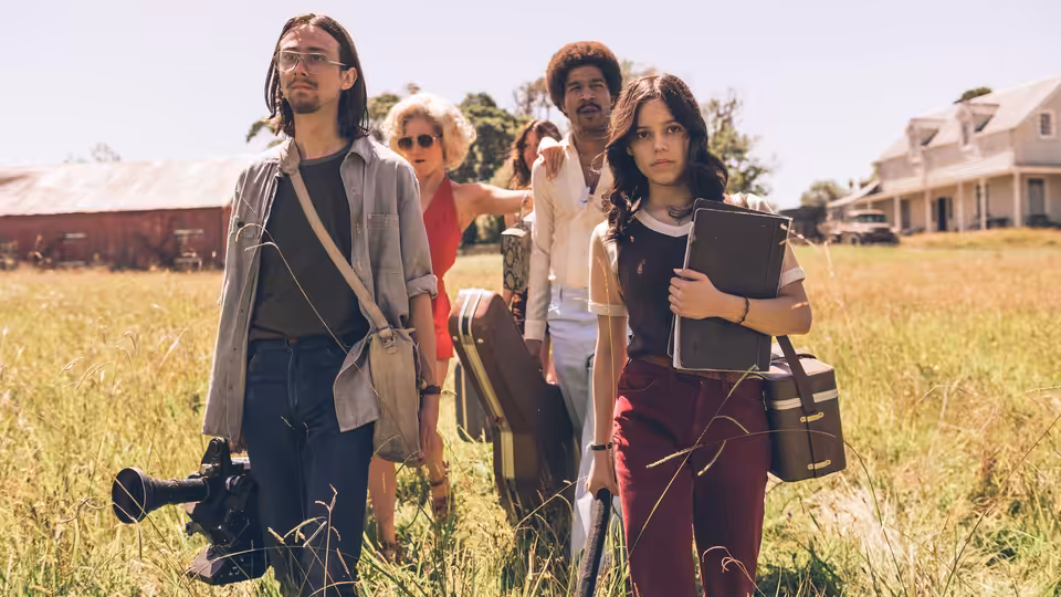 Four people walk through a grassy field carrying luggage and a camera