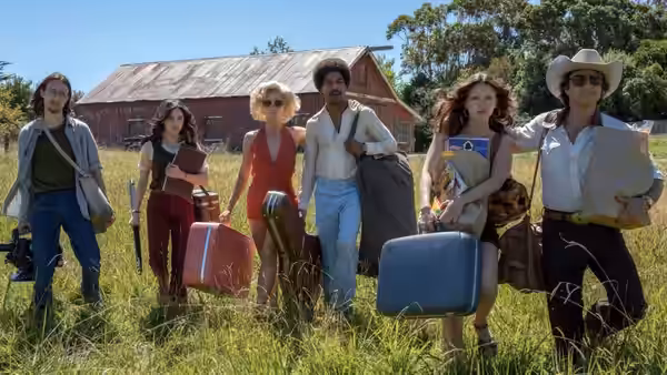 Six people stand in a field holding suitcases in front of a barn
