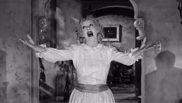 Baby Jane Hudson (Bette Davis) stands with arms outstretched in a parlor