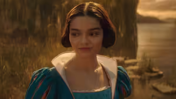 Rachel Zegler as Snow White stands outdoors by a lake wearing a blue dress