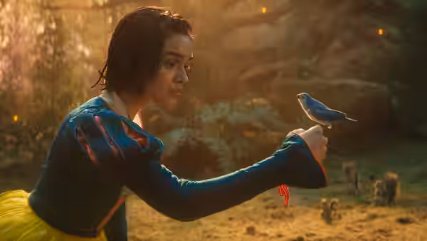 Rachel Zegler as Snow White extends her hand toward a blue bird in a forest